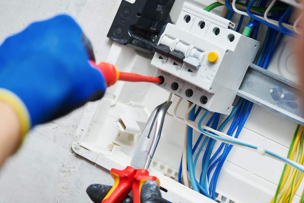 Person wearing blue gloves uses tools to work on an electrical circuit box with colored wires and switches.