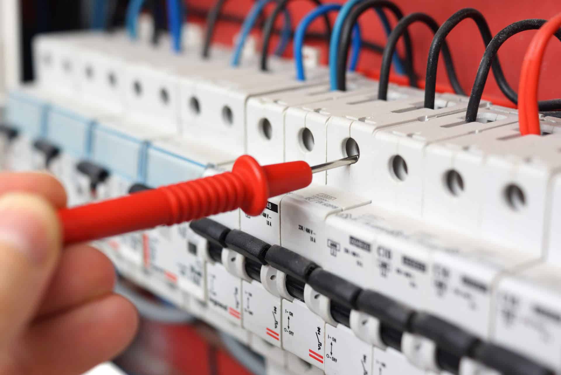 Close-up of a hand holding a red electrical probe testing a row of circuit breakers in an electrical panel.