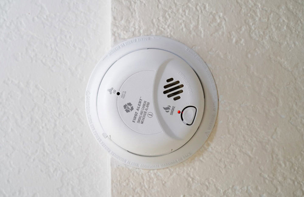 Wall-mounted smoke detector with a test button and a vented cover, installed on a textured white ceiling.