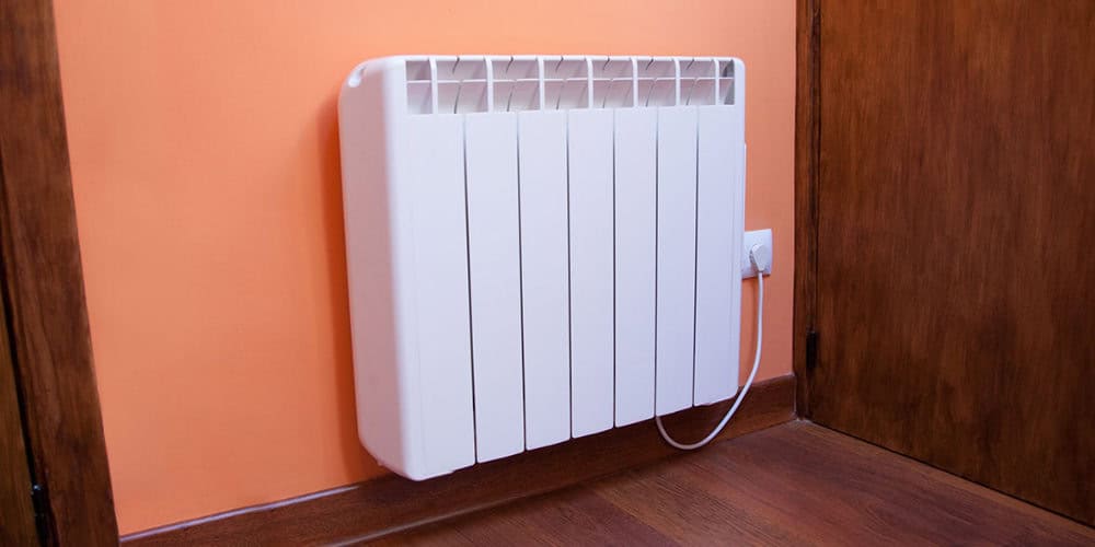 A white electric heater is mounted on an orange wall, plugged into a nearby socket. The floor is wooden.